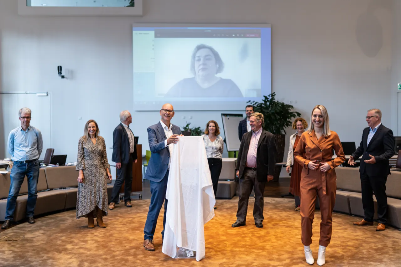 Onthulling in een raadzaal, met een spreker op het scherm
